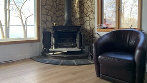 A room with a stone fireplace, a leather chair, and large windows with a view of trees. - Lakeside Sunset Cottage & Hot tub (Alburgh)