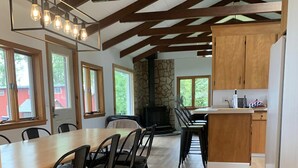 A spacious dining area with a long wooden table and chairs, a stone fireplace, and a kitchen area with wooden cabinets. - Lakeside Sunset Cottage & Hot tub (Alburgh)