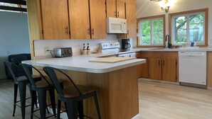 A kitchen with wooden cabinets, a white countertop, a central island with bar stools, a microwave, a coffee maker, and a dishwasher. - Lakeside Sunset Cottage & Hot tub (Alburgh)