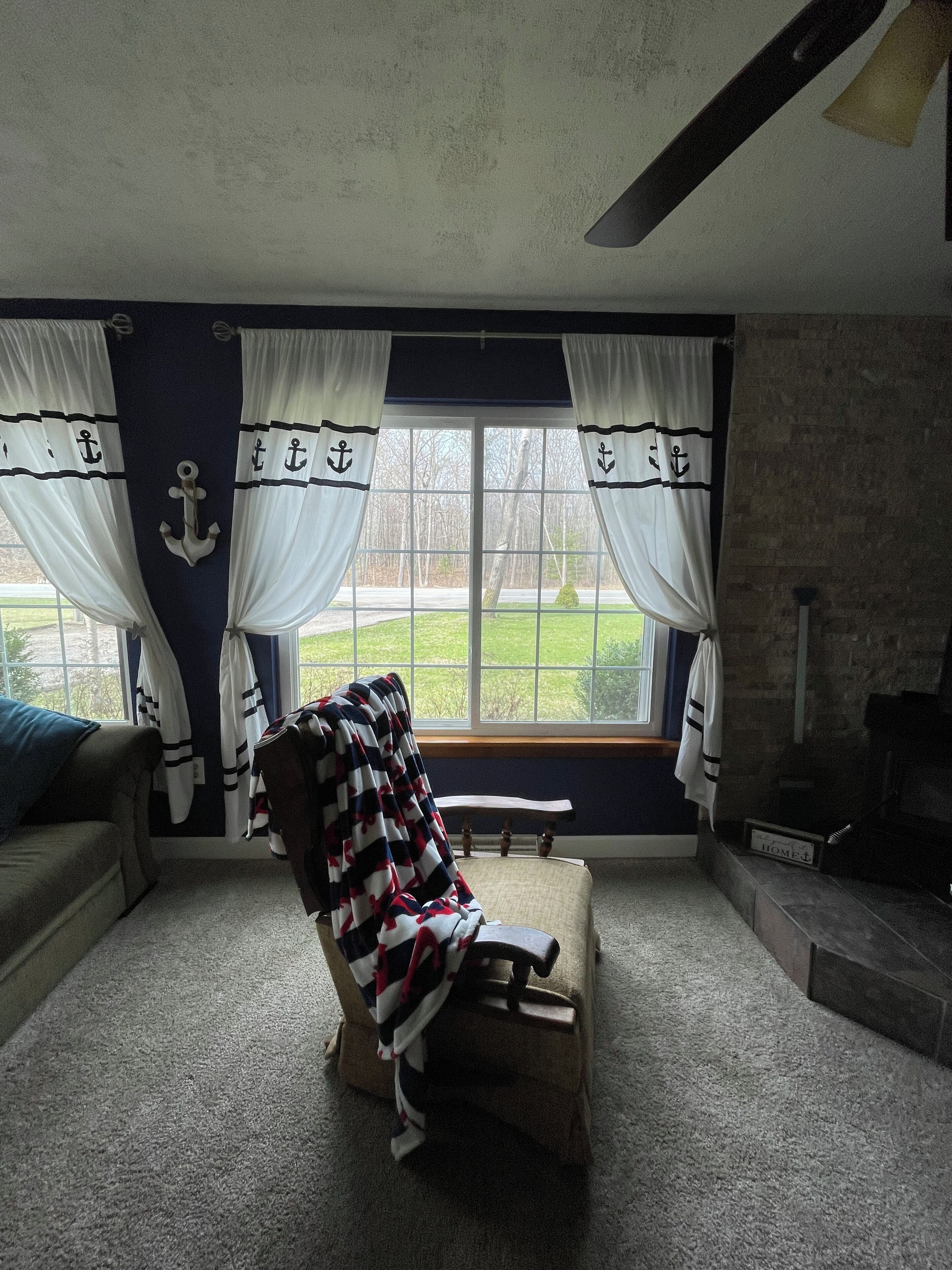 A room with a large window, a chair with a blanket, a fireplace, and curtains with anchor designs.