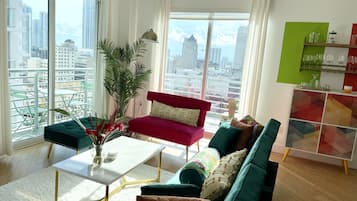 A modern living room with a red sofa, teal armchairs, and a white coffee table. A large plant is in the corner, and there's a view of the city through large windows.