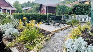 A garden patio with a table and chairs, surrounded by various plants and a white fence.
