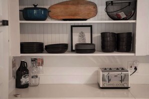 Une cuisine avec des meubles blancs, un pot bleu, une planche à découper en bois, des assiettes noires, un tableau encadré, un grille-pain et une bouteille de kombucha.