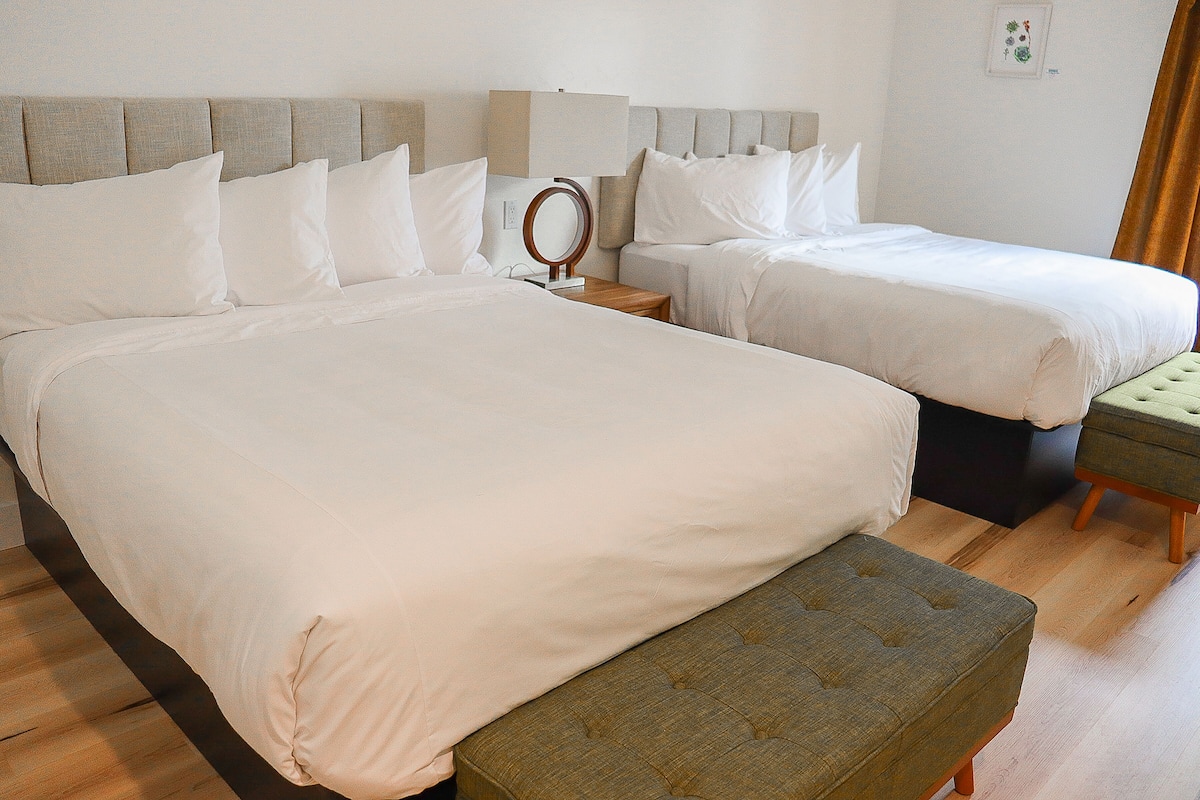 Une chambre d’hôtel avec deux lits, une table de chevet avec une lampe, un petit miroir rond et un pouf vert.