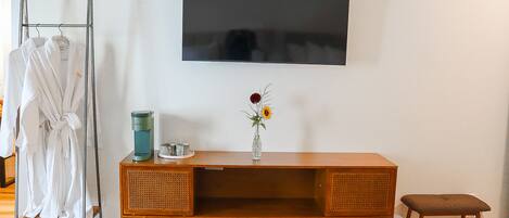 A room with a wooden cabinet, a television mounted on the wall, a small stool, and a rack with white robes.