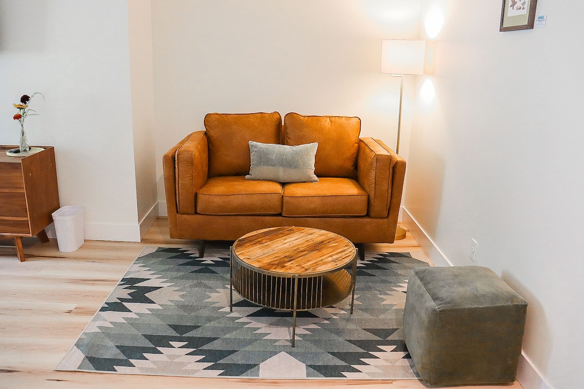 Un salon avec un canapé marron, une table basse ronde en bois, un pouf gris et un tapis à motifs géométriques.