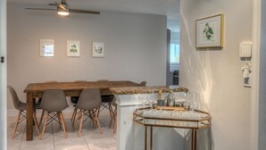 Un coin repas comprenant une table en bois, quatre chaises, un ventilateur de plafond et un chariot à boissons avec des verres.