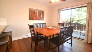 Un comedor con mesa y sillas de madera, un ventilador de techo, un cuadro en la pared y un ventanal corredizo que da a un espacio exterior.