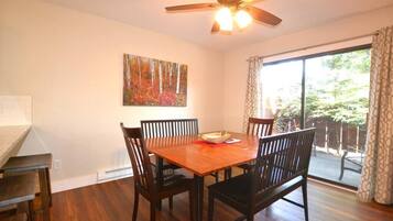Un coin repas avec une table et des chaises en bois, un ventilateur de plafond, un tableau accroché au mur et une porte-fenêtre coulissante donnant sur un espace extérieur.