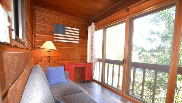 Interior acogedor de una cabaña con una pared de madera, un escritorio rojo, un cojín azul y un ventanal con vistas a árboles.