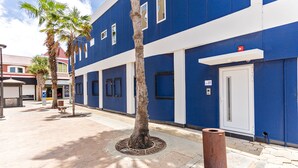Een blauw gebouw met een witte deur, palmbomen en een afvalbak op de stoep.