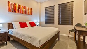 Een slaapkamer met een houten bed, wit beddengoed, rode kussens, een bureau en twee ramen met zonwering.