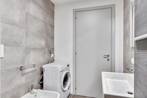 A modern bathroom with grey tiles, a washing machine, a sink, and a door. - Appartamento Mango - Affitti Brevi Italia (Lecco)