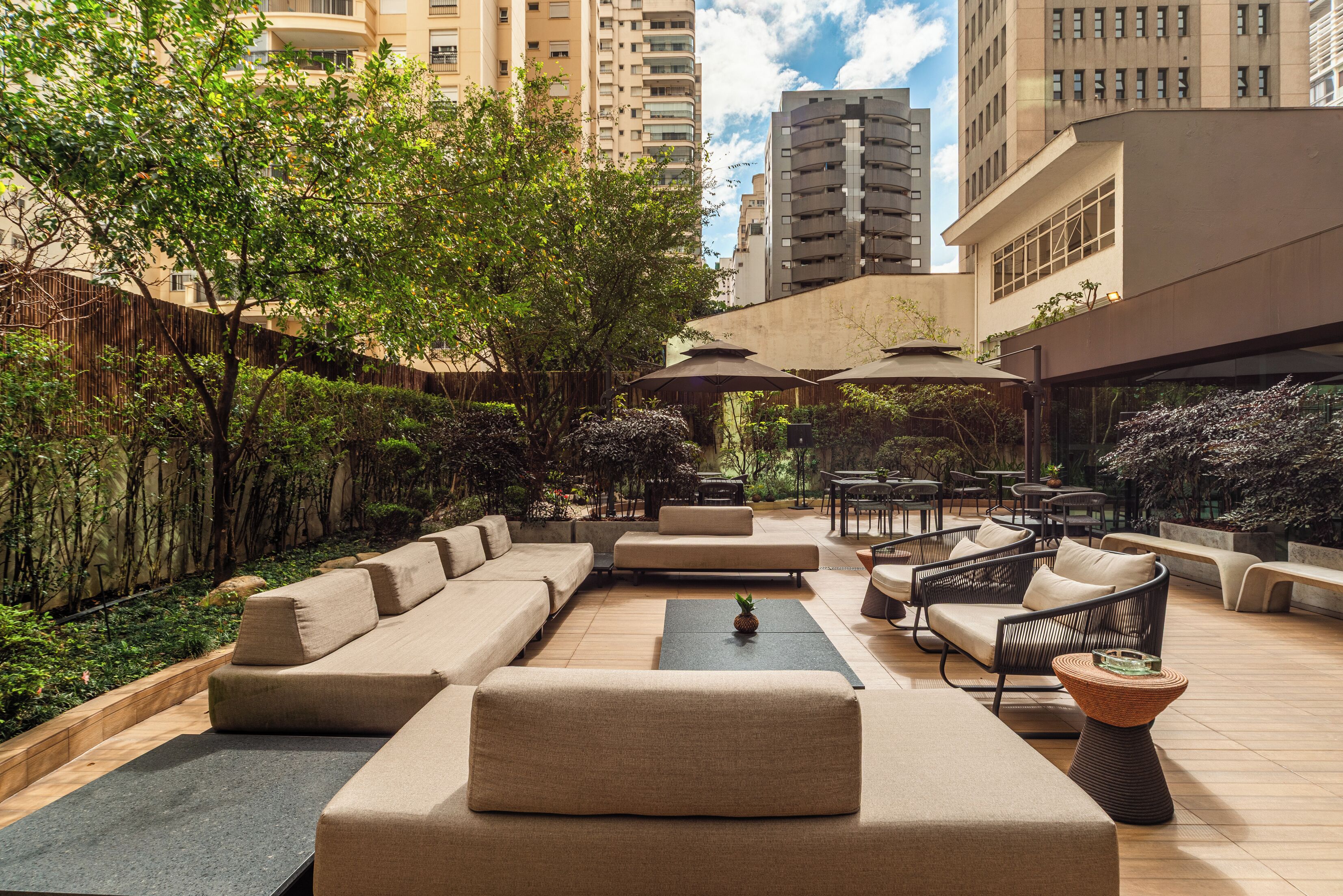 Un espace extérieur aménagé avec des canapés, des tables et des parasols, entouré de verdure et de bâtiments imposants.