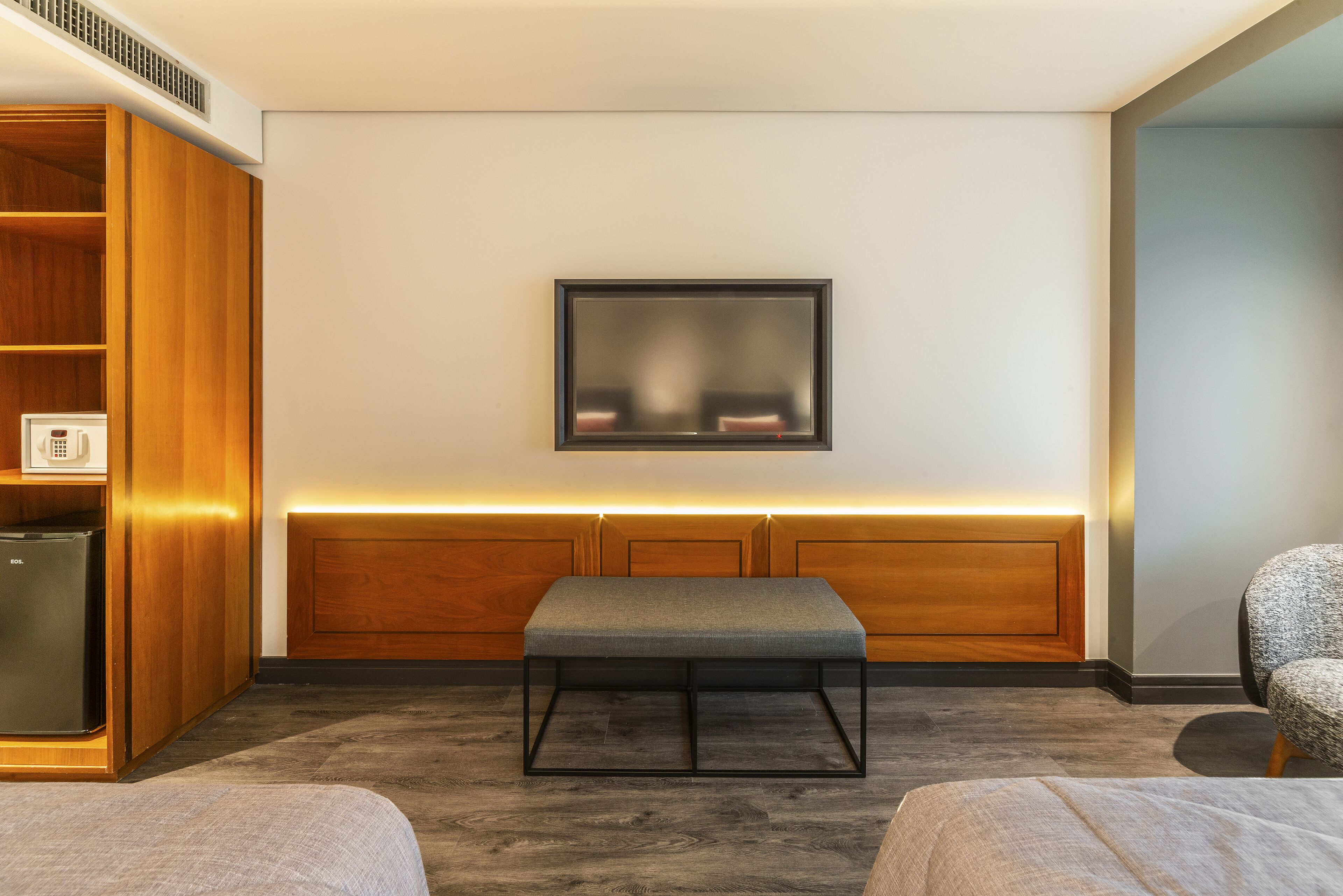 Una habitación de hotel moderna con un armario de madera, un banco, un televisor montado en la pared y una zona de estar.