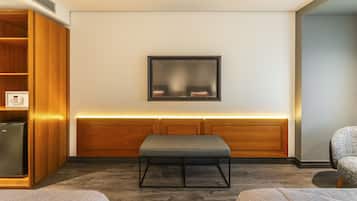 A modern hotel room with a wooden wardrobe, a bench, a wall-mounted TV, and a seating area.