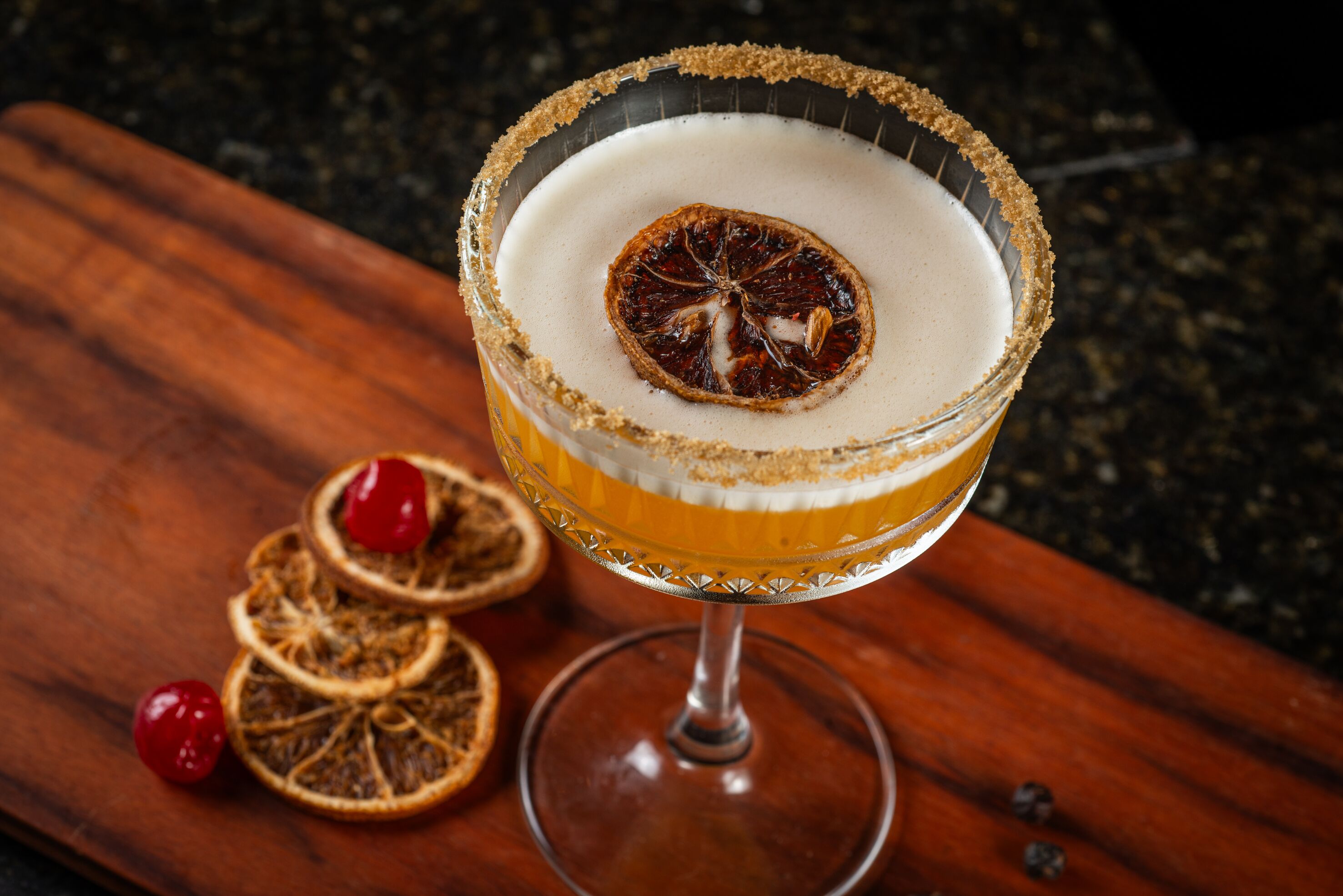 Um cocktail num copo com borda salgada e rodela de laranja desidratada, sobre uma superfície de madeira com rodelas de laranja secas e cereja.