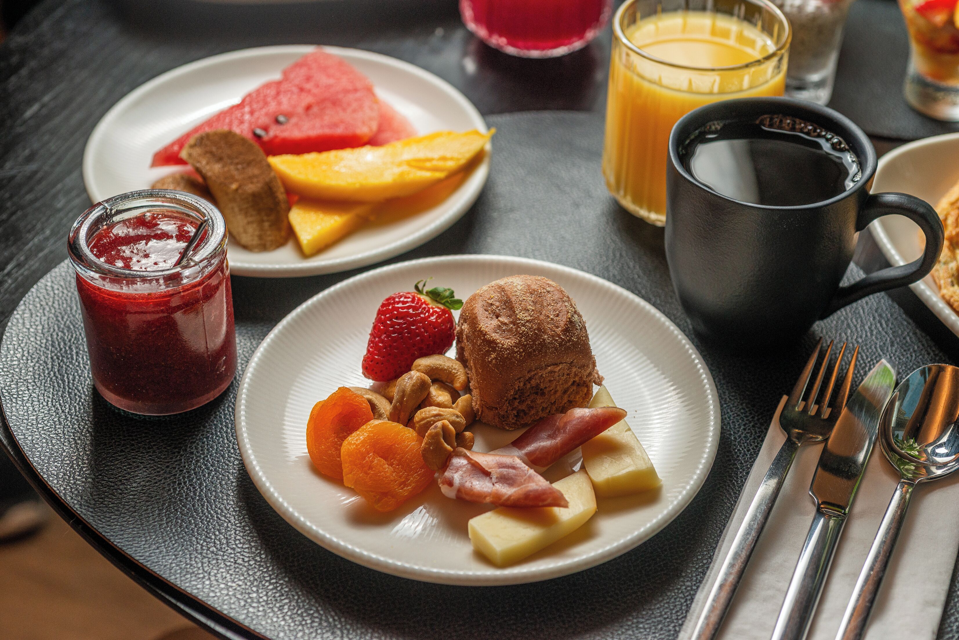 Desayuno con platos de comida, un vaso de jugo de naranja, un frasco de mermelada y una taza negra.