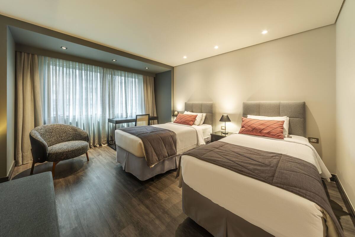 Une chambre d’hôtel avec deux lits, une chaise, un bureau et une fenêtre avec des rideaux.