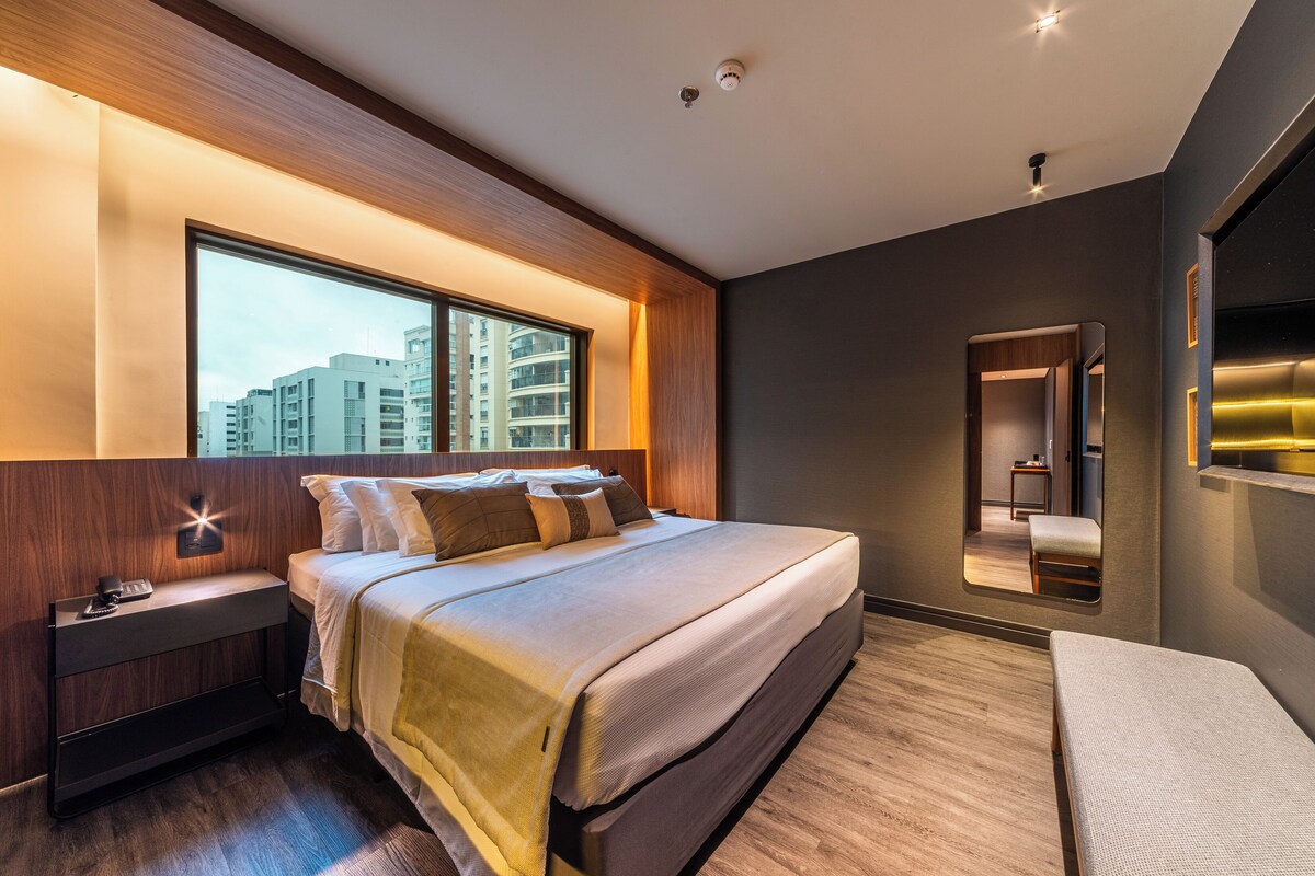 A modern bedroom with a large window, a bedside table, a bed, a mirror, and a bench.