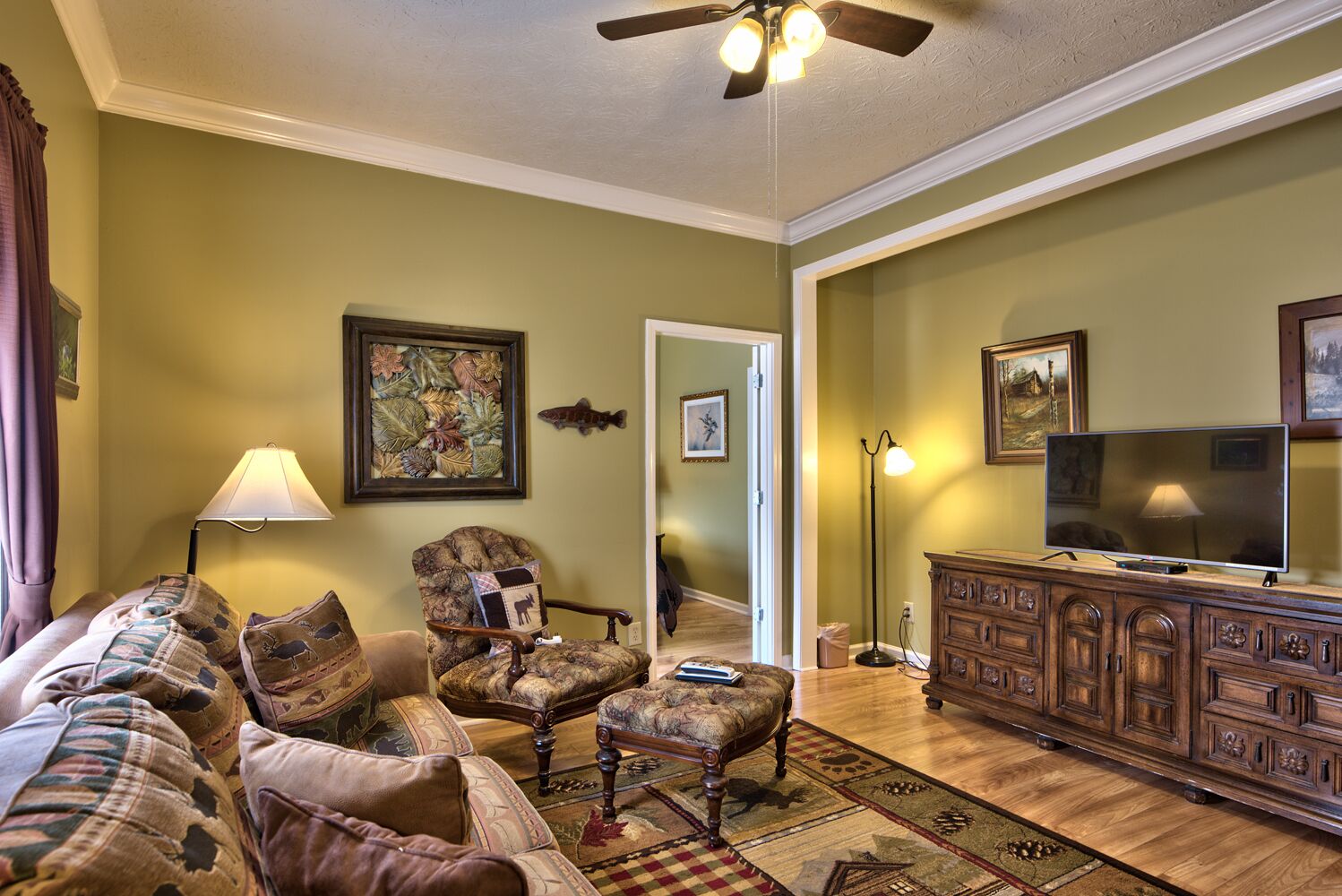A living room with a sofa, armchair, ottoman, wooden cabinet, television, and a ceiling fan.