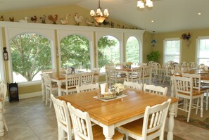 A dining area with multiple tables and chairs, large windows with views of trees, and a ceiling with decorative lights.