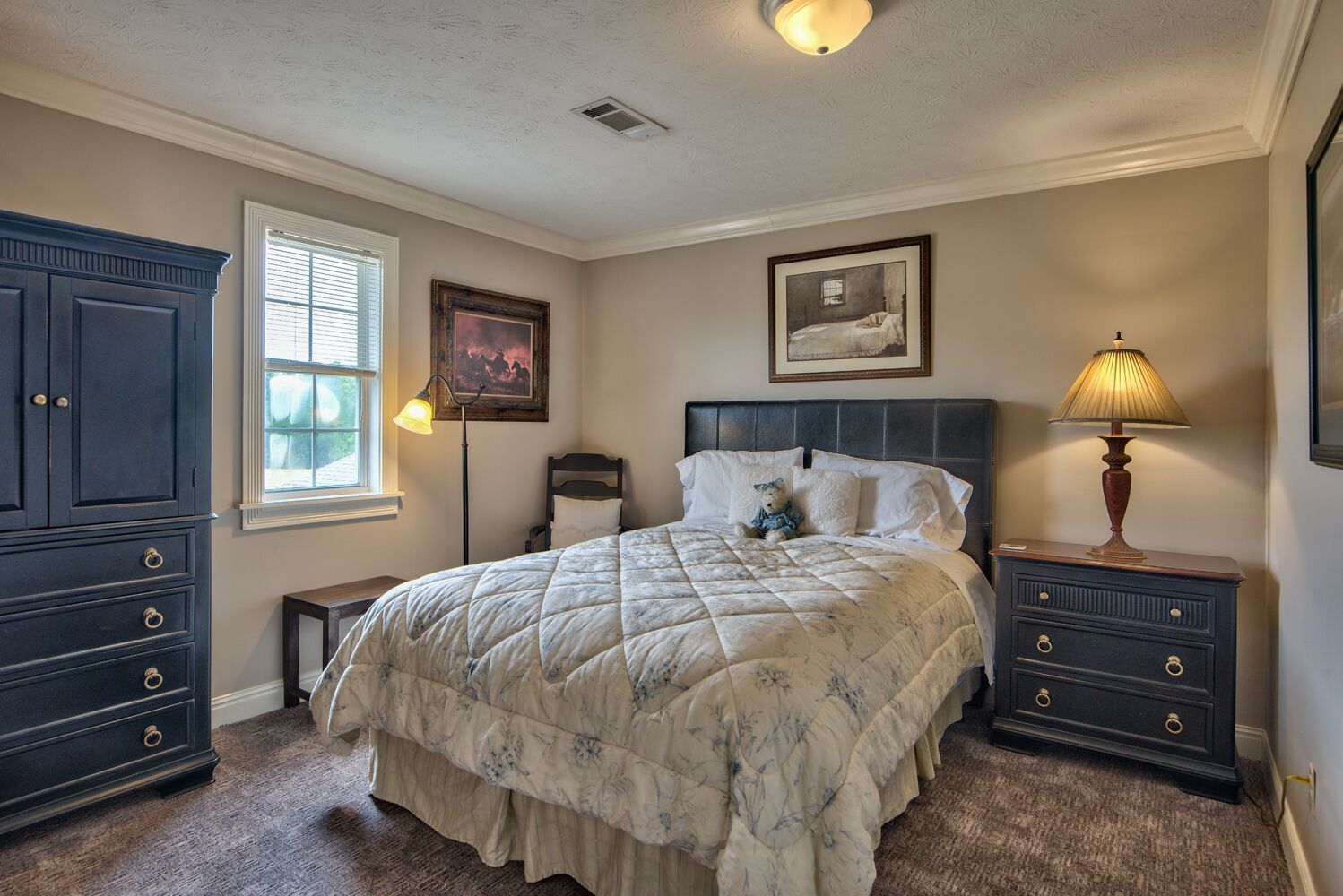 A bedroom with a bed, a dresser, a nightstand, a lamp, and two framed pictures on the wall.