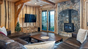 A cozy living room with a stone fireplace, leather sofas, a wooden coffee table, and a television mounted on the wall. - One-of-A-Kind Log Home in the Heart of the White Mountains NH (Intervale)