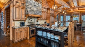 A spacious log cabin interior with a large kitchen area featuring wooden cabinets, a stone fireplace, and a dining area with wooden chairs. - One-of-A-Kind Log Home in the Heart of the White Mountains NH (Intervale)