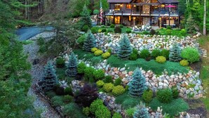A log cabin with a stone foundation, surrounded by a landscaped garden with various shrubs and trees, and a small stream running through the property. - One-of-A-Kind Log Home in the Heart of the White Mountains NH (Intervale)