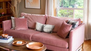 Un sofá rosa con varios cojines, una mesa de centro de madera con cuencos de cobre y una estantería con adornos.