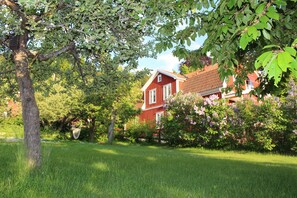 Une maison rouge avec des encadrements de fenêtres blancs, entourée d’une pelouse verdoyante et luxuriante et d’arbres.