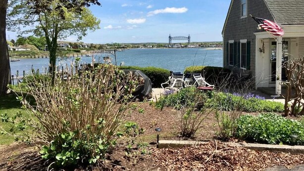 Een huis met uitzicht op een brug over het water, een tuin met planten en een terras met stoelen.