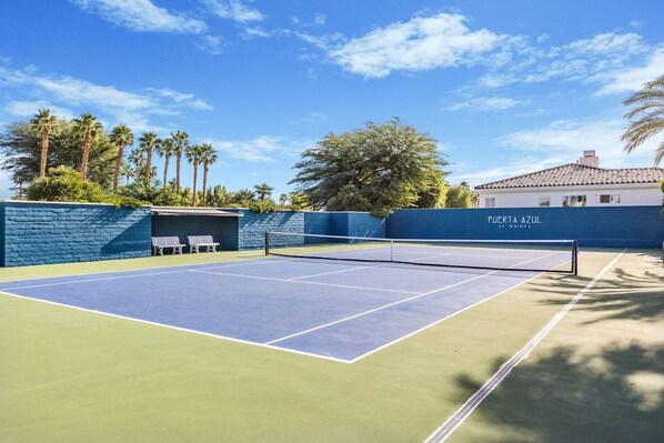 A tennis court with a blue and green surface, surrounded by a blue wall with a white bench, palm trees, and a building with 'PUERTA AZUL LA QUINTA' written on it.
