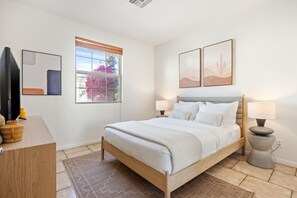 A bedroom with a bed, two bedside lamps, a window with a view of pink flowers, and a wall-mounted TV. - Gated Oasis near Festivals | Pools & Spa Included (La Quinta)