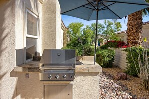 An outdoor grill area with a stone wall, a window, and a patio umbrella. - Gated Oasis near Festivals | Pools & Spa Included (La Quinta)