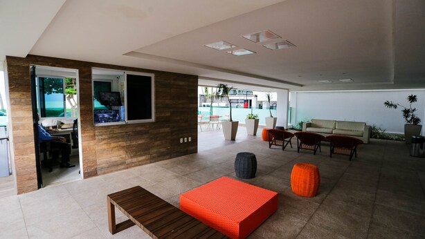 A spacious living area with a wooden bench, a red cushioned seating area, and a view of a pool through large windows. - Apart beira mar Cabo Branco by Carpediem (João Pessoa)