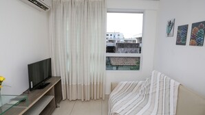 A room with a television on a wooden stand, a window with curtains, a sofa with a striped blanket, and artwork on the wall. - Apart beira mar Cabo Branco by Carpediem (João Pessoa)