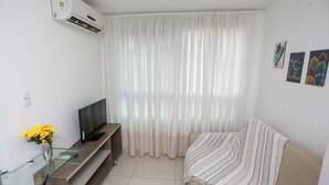 A living room with a TV on a wooden stand, a glass table with a vase of flowers, a sofa with a striped blanket, and a large window with sheer curtains. - Apart beira mar Cabo Branco by Carpediem (João Pessoa)