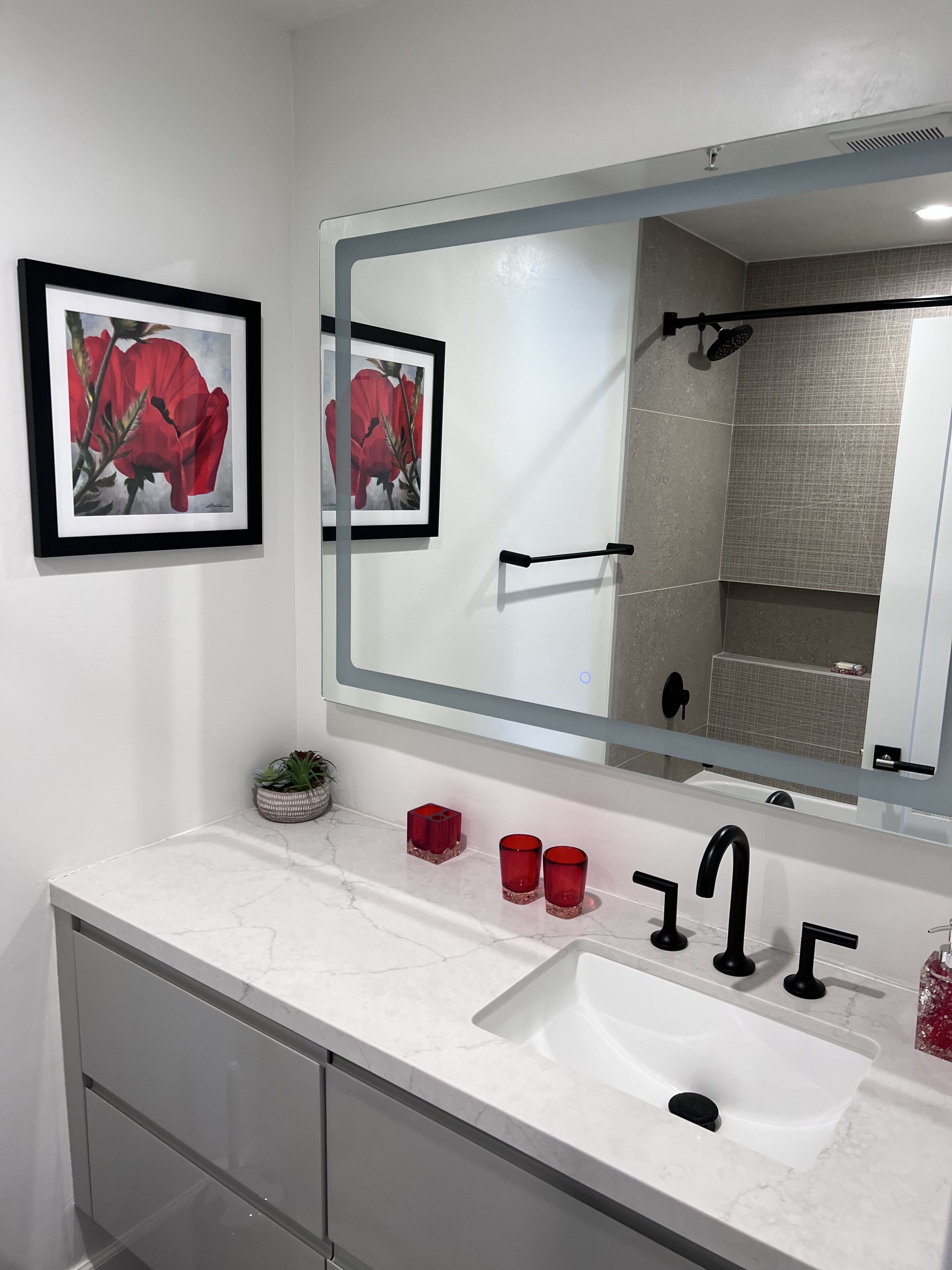 A bathroom with a white marble sink, black faucet, and a large mirror. There are two red candle holders and a small potted plant on the counter.
