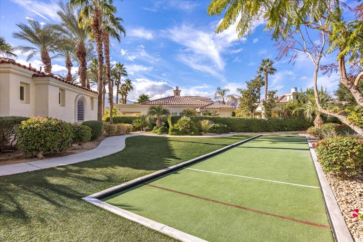A croquet field with a clear path, surrounded by well-maintained hedges and palm trees.