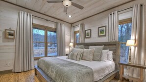 A bedroom with a large bed, wooden headboard, and a ceiling fan. There are two windows with curtains, a nightstand with a lamp, and a framed picture on the wall. - Hot Tub- Modern Luxury -5 Bdrm-4 Kings- Pool Table (Blue Ridge)