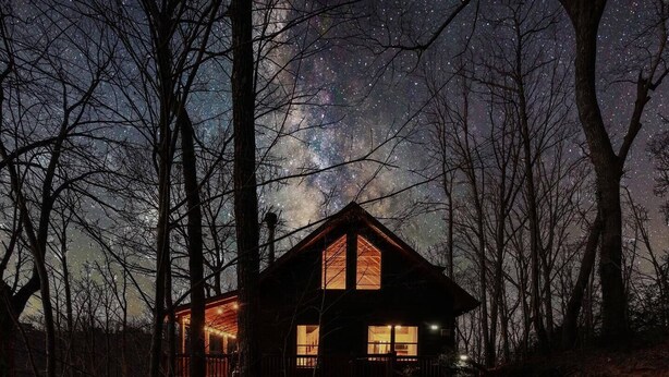 A cabin with a large window, surrounded by trees, under a starry night sky. - Sunset-View 3BR Blue Ridge Cabin • Hot Tub (Cherry Log)