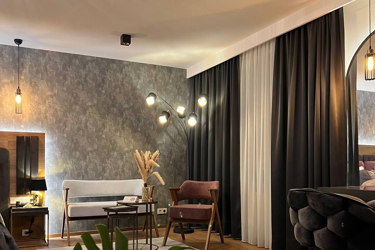 A modern living room with a sofa, a dining table, and chairs. There is a large window with curtains, a plant, and a lamp.