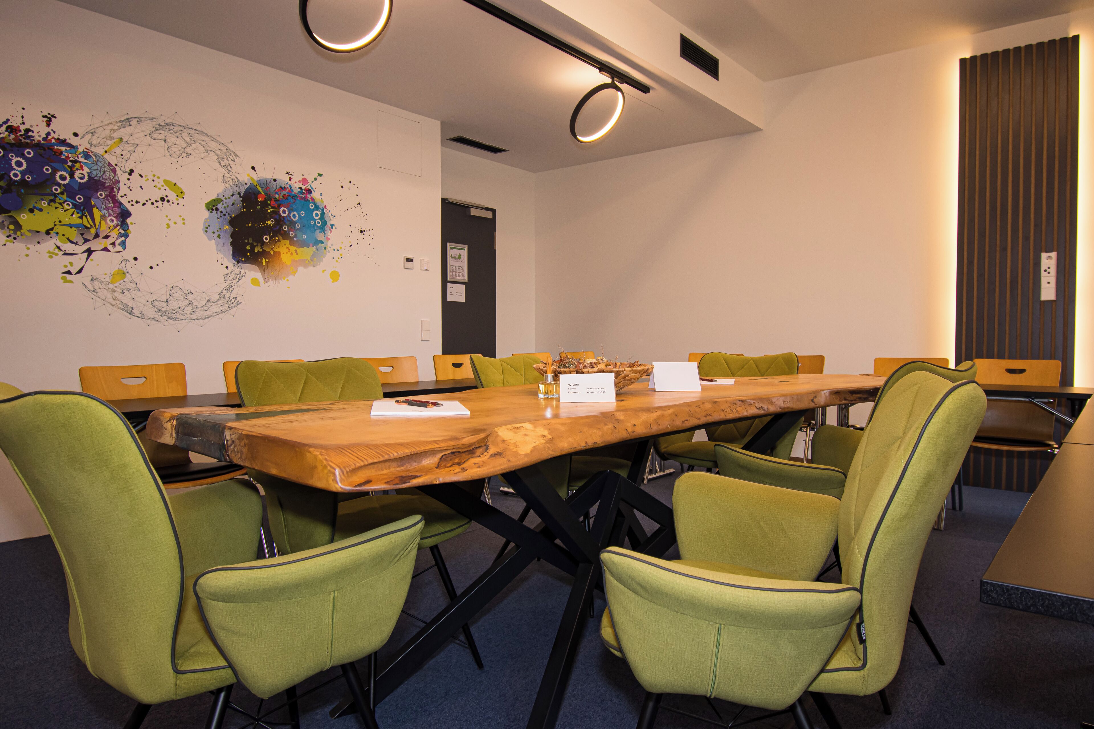 Une salle de conférence avec une table en bois, des chaises recouvertes d’un tissu vert et une œuvre d’art abstraite et colorée accrochée au mur.