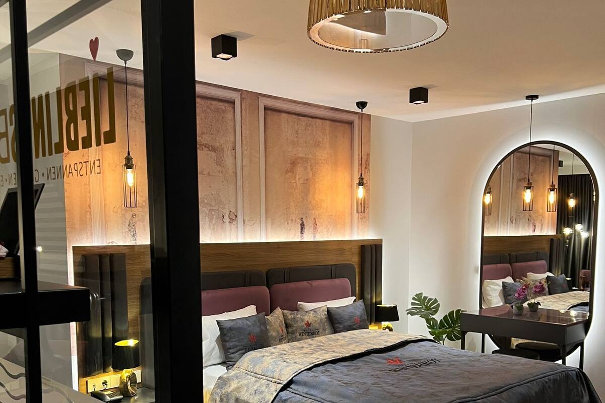 A modern bedroom with a large bed, wooden headboard, and a mirror above the bed. There is a small desk with a chair and a potted plant.