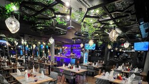 A restaurant with multiple tables set for dining, a bar area in the background, and hanging plants. - Hotel Winterrot (Karlsruhe)