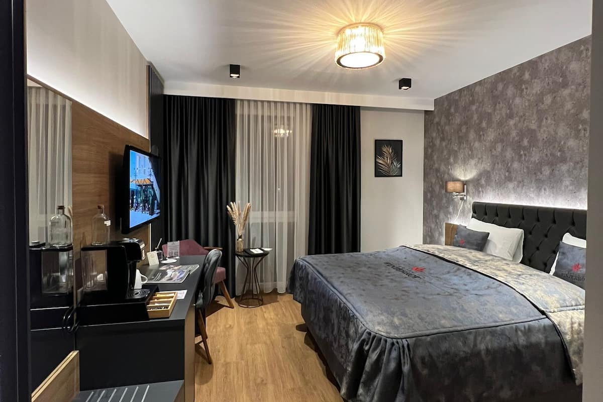 A modern hotel room with a large bed, a desk with a coffee machine, a TV, and a minibar.