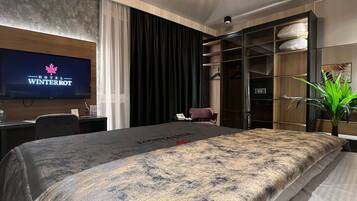 Ein modernes Hotelzimmer mit einem großen Bett, einem Fernseher, der 'Hotel Winterrot' zeigt, einem Schreibtisch mit einem Laptop, einem Kleiderschrank und einer Topfpflanze.