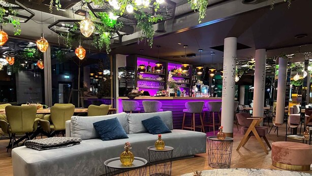 A modern lounge area with a grey sofa, round tables, and hanging plants. - Hotel Winterrot (Karlsruhe)
